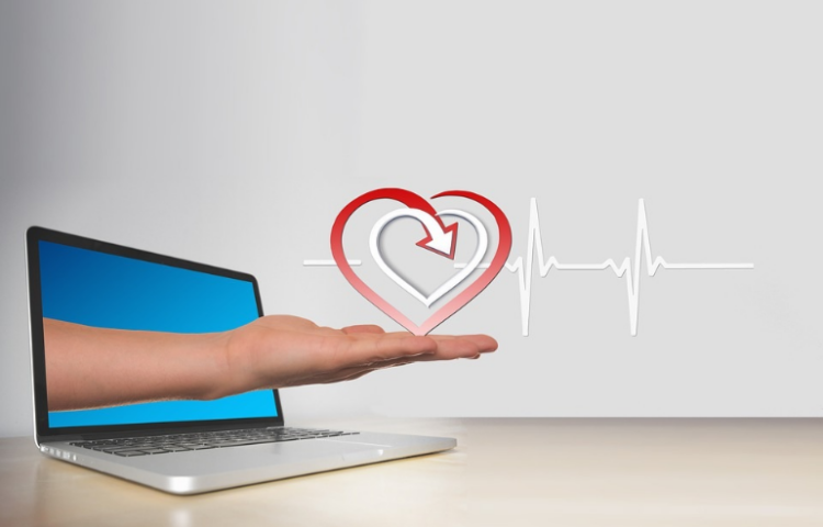 An illustration of a hand holding a heart emerging from a laptop, illustrating the debate of telehealth vs in-person rehab.