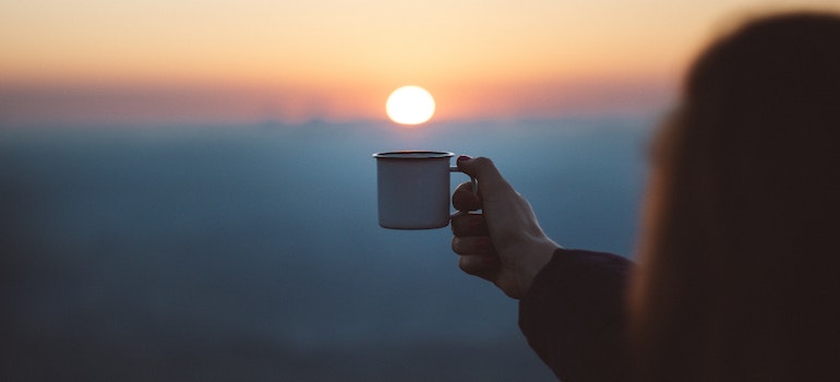 A person holding a cup extending their hand toward the sun